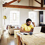 A guy wearing a yellow t-shirt works from home using his dining room table as a desk