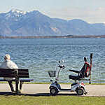 Back view of older man sitting on a bench on the waterfront with an electric wheelchair by his side.